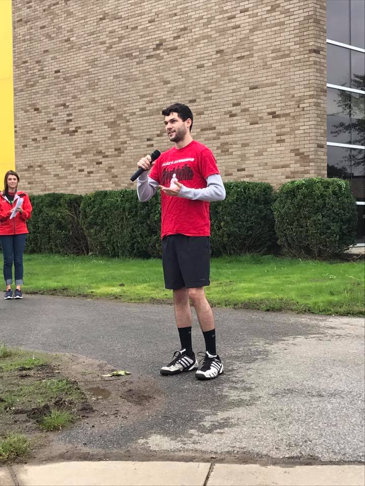 Alex, holding a microphone, addressing a group and wearing a red shirt and black shorts