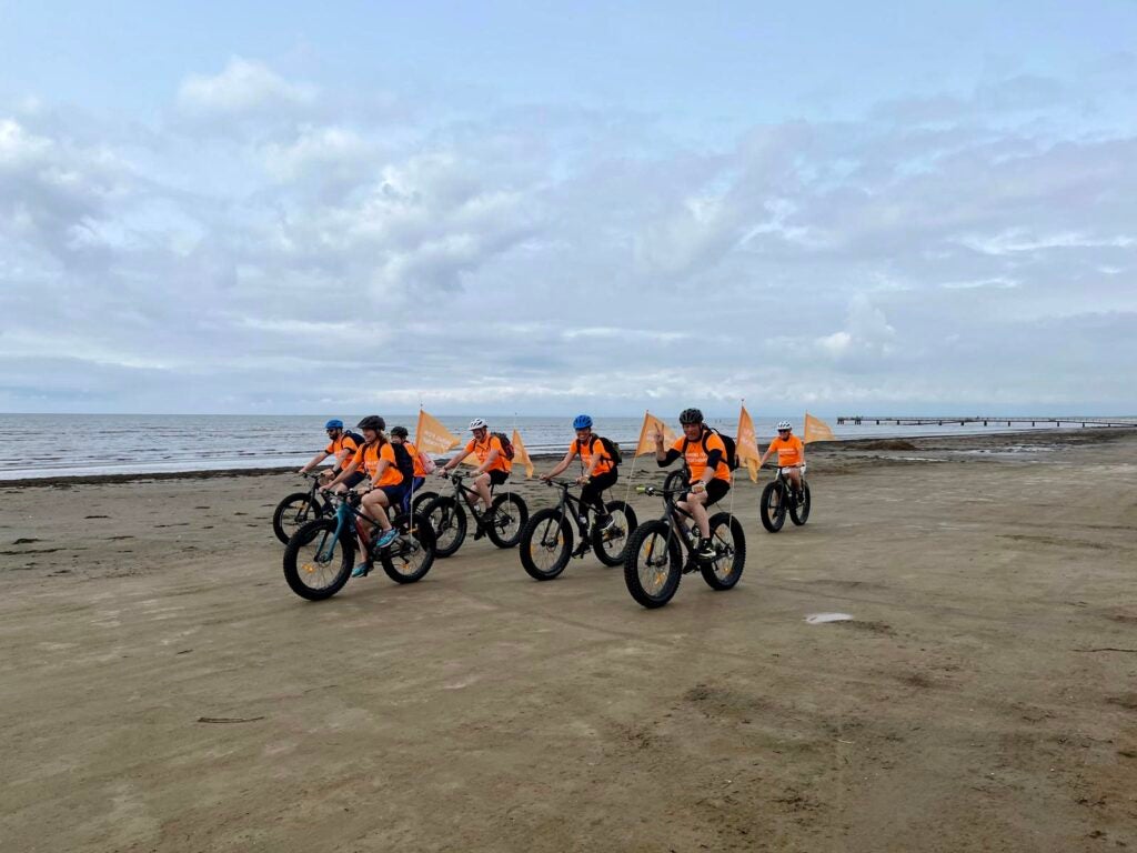 Group of PTC employees in orange shirts, riding their bikes on the beach in support of the De Duchenne 40.