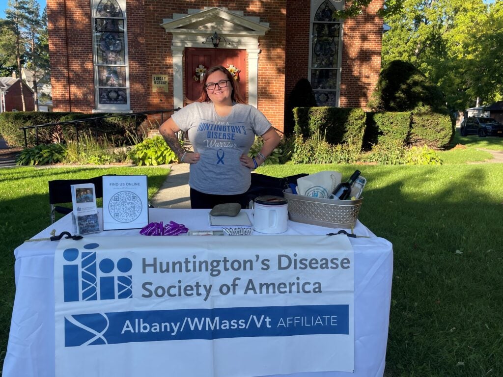 Jenna, standing behind a table wearing a shirt that says "I am a Huntington's Disease Warrior"