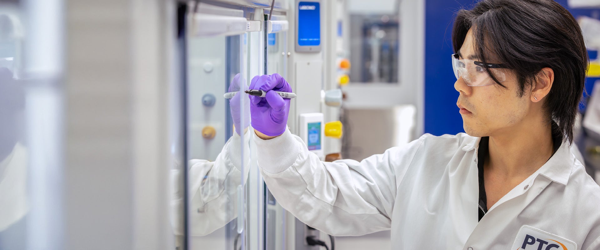 Scientist in white lab coat and purple gloves, wearing eye protection, writing on glass with a marker in the lab